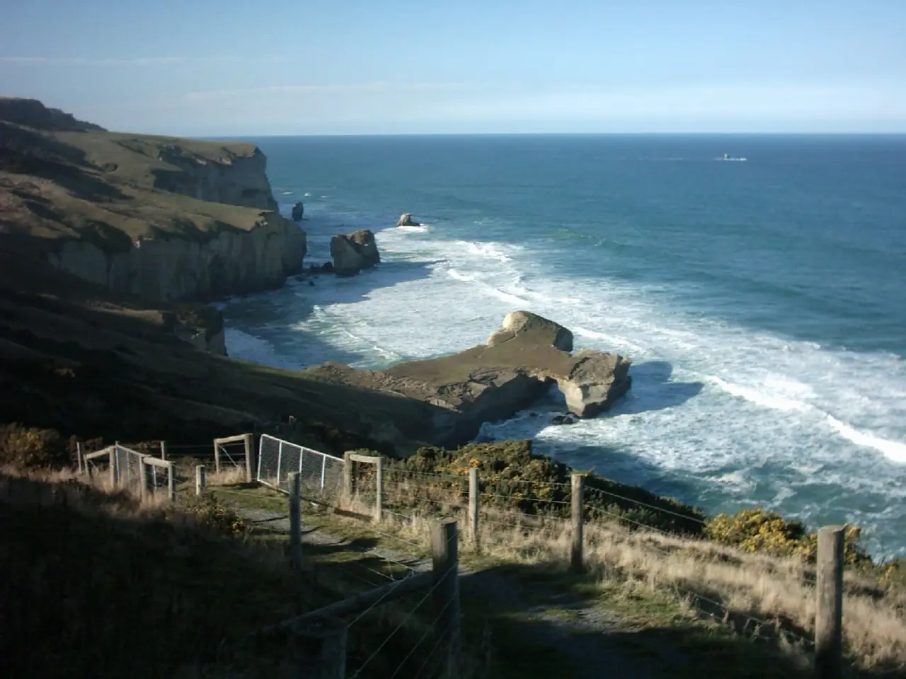 ชายฝั่งหินและทะเลที่ Tunnel Beach มองจากทางเดินที่มีรั้วและประตู ภาพจาก Wikimedia Commons ผู้ถ่าย Ingolfson สัญญา public domain เป็นภาพเก่าไม่ระบุปีที่ถ่าย สภาพทางในภาพอาจไม่ตรงกับปัจจุบัน