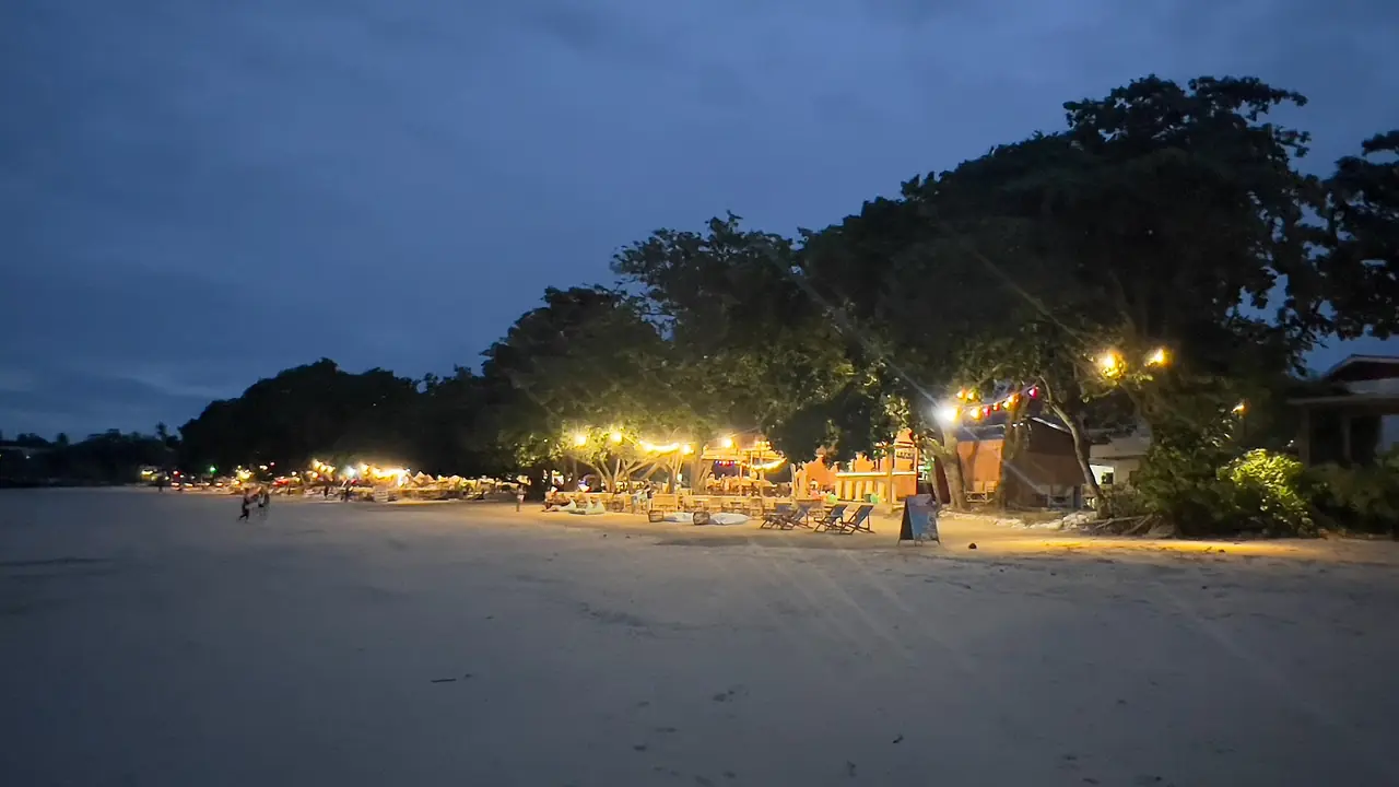หาดคลองม่วงตอนค่ำ มีไฟสีส้มจากร้านริมหาดเรียงอยู่ใต้ต้นไม้ ภาพจาก Wikimedia Commons ผู้ถ่าย Stefan Fussan สัญญา CC BY-SA 4.0 ไม่ระบุปีที่ถ่าย บล็อก homeiswhereyourbagis ว่าเส้นทางขับรถจากอ่าวนางผ่านคลองม่วง เป็นภาพประกอบของบริเวณใกล้ทาง ไม่ใช่ตัวเขา
