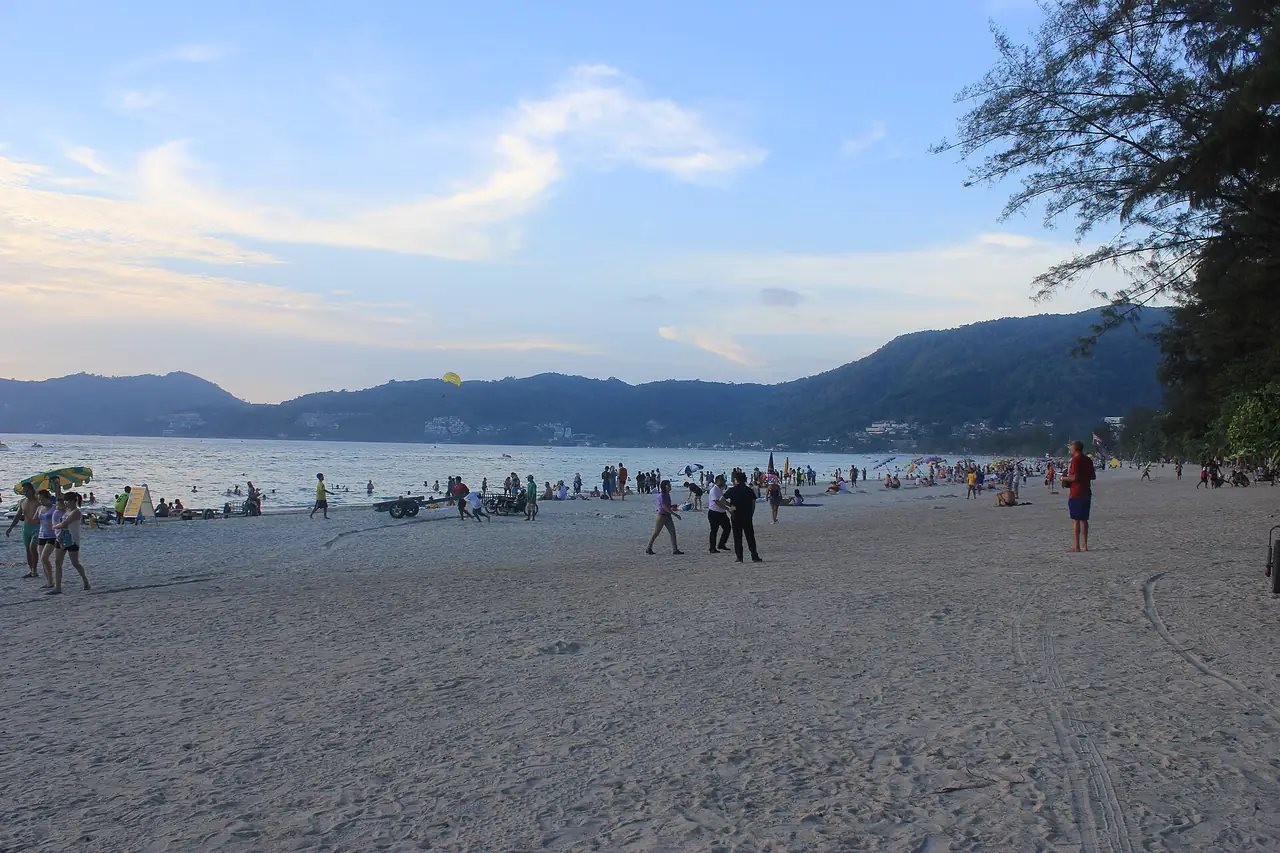 หาดป่าตองช่วงเย็นมีคนเดินและเล่นน้ำ ภูเขาเป็นฉากหลังและมีร่มชูชีพลอยอยู่ไกล ๆ ภาพจาก Wikimedia Commons ผู้ถ่าย scott paul riefler สัญญา CC BY-SA 4.0 ไม่ระบุปีที่ถ่าย เป็นภาพประกอบ