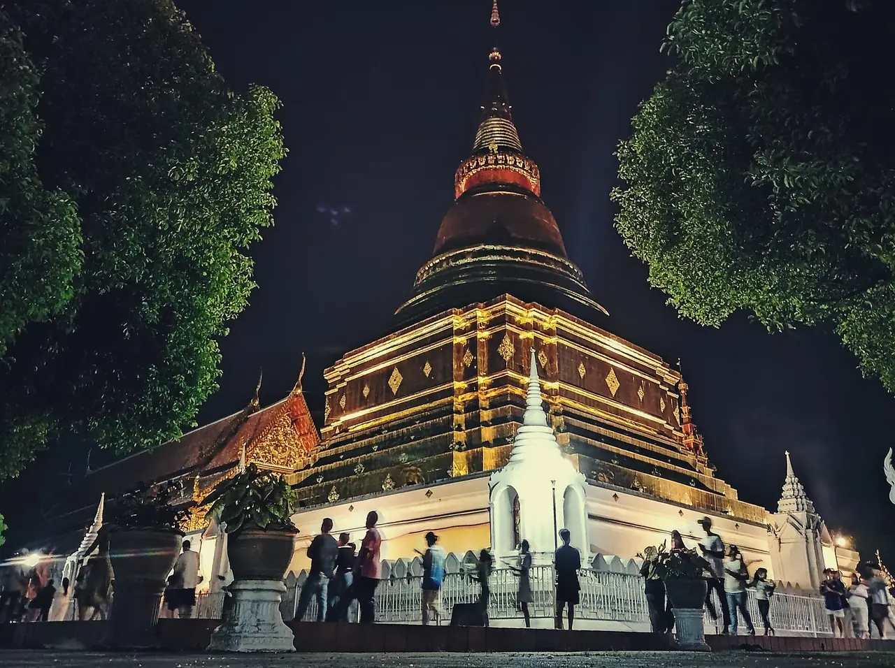 พระบรมธาตุดอนเต้าตอนกลางคืนที่เปิดไฟส่ององค์เจดีย์ มีผู้คนยืนชมรอบฐาน ภาพของ GAYATANA จาก Wikimedia Commons สัญญา CC BY-SA 4.0 ไฟล์ตั้งชื่อว่าเป็นงานมาฆบูชา ปีที่ถ่ายไม่ระบุที่เราตรวจ