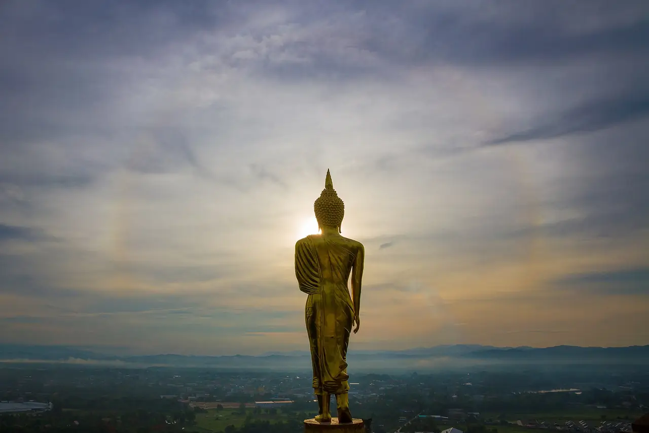 พระพุทธรูปสีทองยืนอยู่ปลายลานมองจากด้านหลัง ดวงอาทิตย์อยู่ตรงหน้าพระและมีวงแสงจาง ๆ ด้านซ้าย ไกลออกไปเป็นเมืองและแนวเขาในหมอกบาง ภาพจาก Wikimedia Commons ผู้ถ่าย Manoonp สัญญา CC BY-SA 4.0 ไม่ลงวันที่ถ่าย เป็นภาพประกอบ