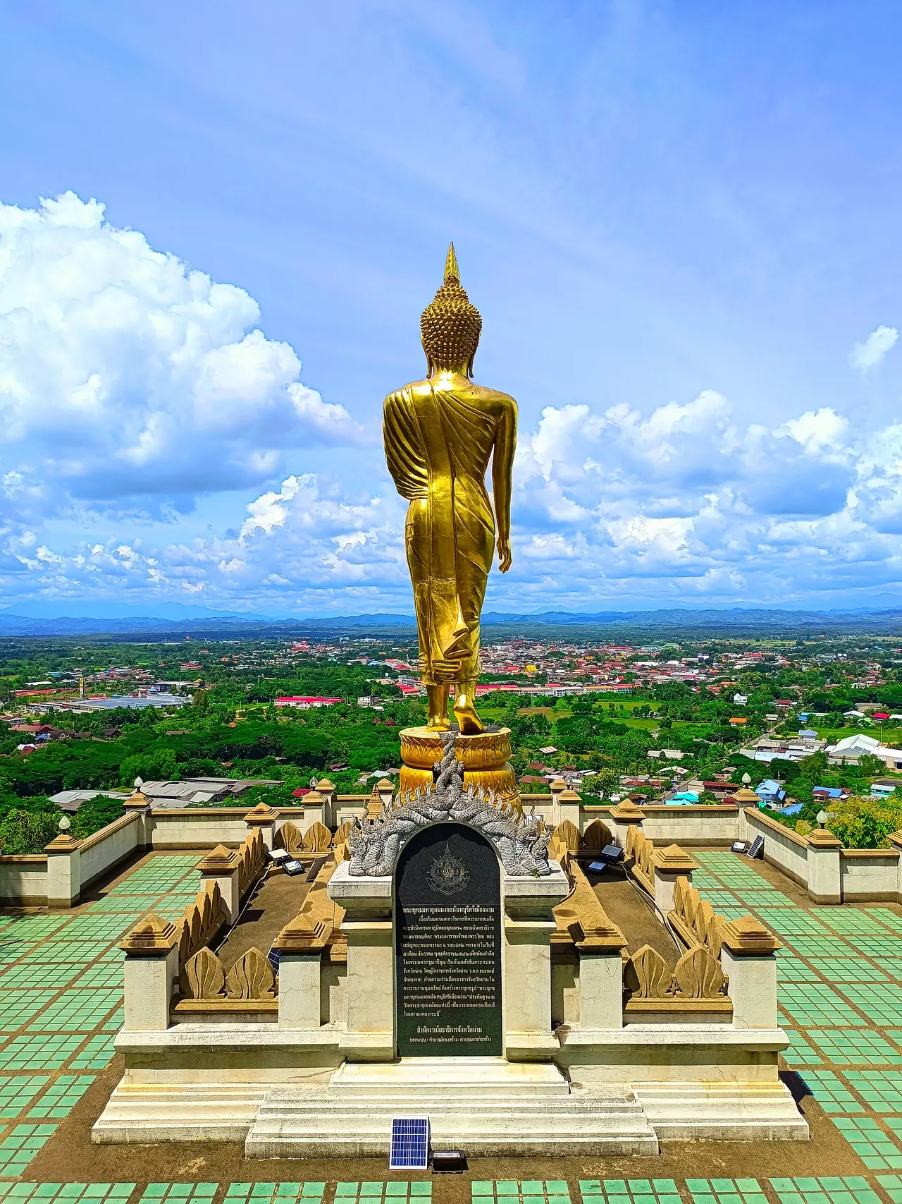 พระพุทธรูปสีทองมองจากด้านหลังตอนท้องฟ้าเมฆมาก มีป้ายแผ่นศิลาเขียนข้อความและรูปพญานาคหรือลายประดับที่ฐาน ด้านหลังเป็นเมืองและแนวเขาไกล ๆ ภาพจาก Wikimedia Commons ผู้ถ่าย Jomis 9 สัญญา CC BY-SA 4.0 ชื่อไฟล์ระบุปี 2022 เราไม่ได้อ่านข้อความบนป้ายจากภาพนี้ เป็นภาพประกอบ