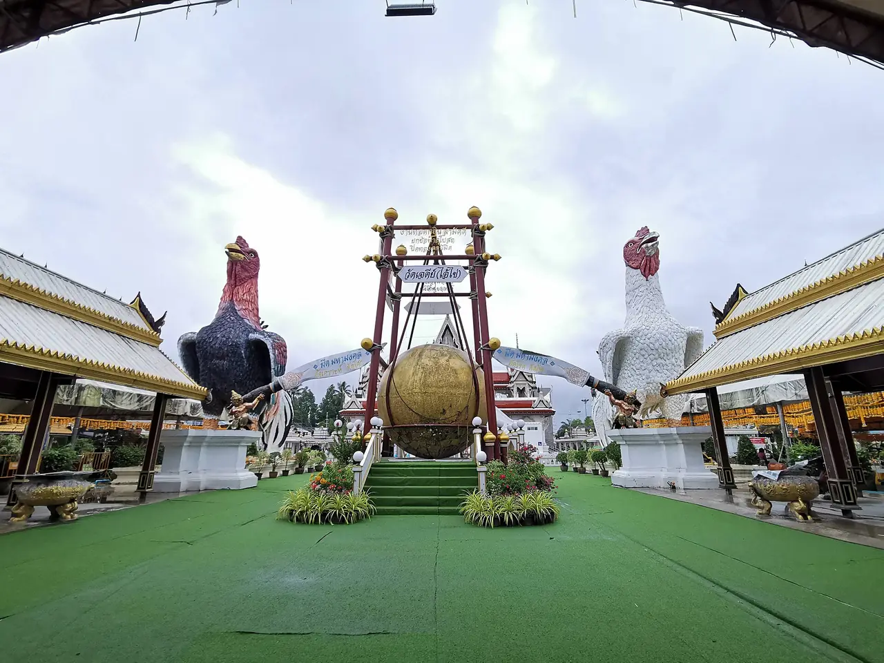 รูปปั้นไก่ชนขนาดใหญ่สองตัวยืนบนแท่นสองฝั่งของลานวัด มีกรอบฆ้องใหญ่ตั้งอยู่ตรงกลาง คำอธิบายไฟล์ระบุว่าเป็นที่วัดเจดีย์ (ไอ้ไข่) อำเภอสิชล ภาพจาก Wikimedia Commons ผู้ถ่าย OraMAAG สัญญา CC BY-SA 4.0 ไม่ระบุปีที่ถ่าย อาจต่างจากปัจจุบัน