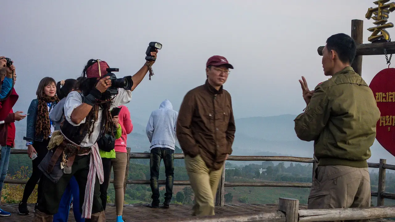 ผู้คนยืนบนลานไม้ริมราวชมวิวยามเช้า ฟ้าเทาฟ้าและหมอกบาง มีป้ายไม้อักษรจีนสีทองและสีแดงอยู่มุมขวาบน ชายถือกล้องแฟนซีเป็นโจรสลัดอยู่ซ้ายกลางภาพ และผู้ชายเสื้อน้ำตาลเดินผ่านกลางภาพ ชื่อไฟล์คือจุดชมวิวหยุนไหล ปาย ภาพจาก Wikimedia Commons ผู้ถ่าย Visions of Domino สัญญา CC BY 2.0 ไม่ระบุวันที่ถ่ายในไฟล์ ป้ายและผู้คนอาจเปลี่ยนไปแล้ว
