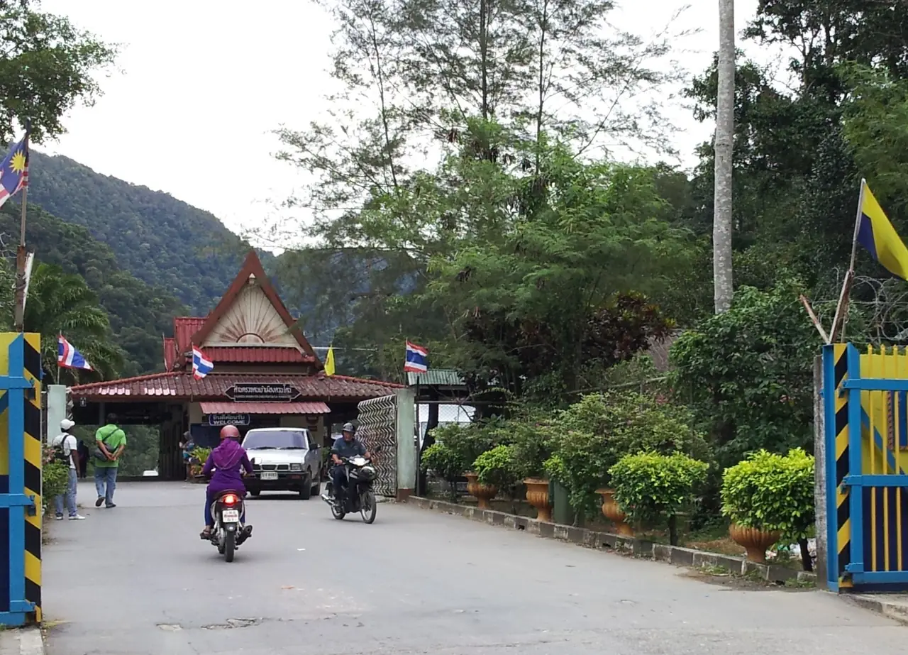 ประตูด่านวังประจัน มองจากฝั่งมาเลเซีย มีธงไทยและธงมาเลเซียและรถจักรยานยนต์ผ่านด่าน ภาพจาก Wikimedia Commons ผู้ถ่าย Slleong สัญญา CC BY-SA 3.0 ไม่ระบุปีที่ถ่าย เป็นภาพประกอบบริเวณชายแดนใกล้ทะเลบัน ไม่ใช่ตัวอุทยาน